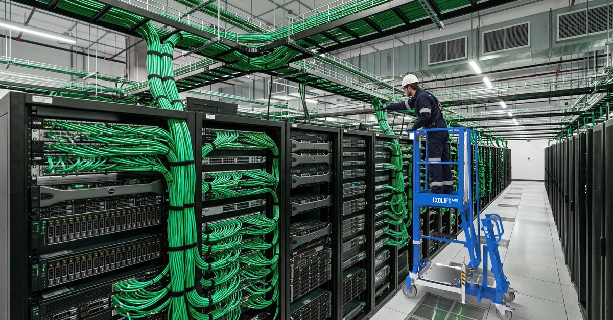 Technician in a white hard hat on a blue IXOLIFT X400 work platform in a data centre aisle, reaching up to bundles of green cables above server racks.