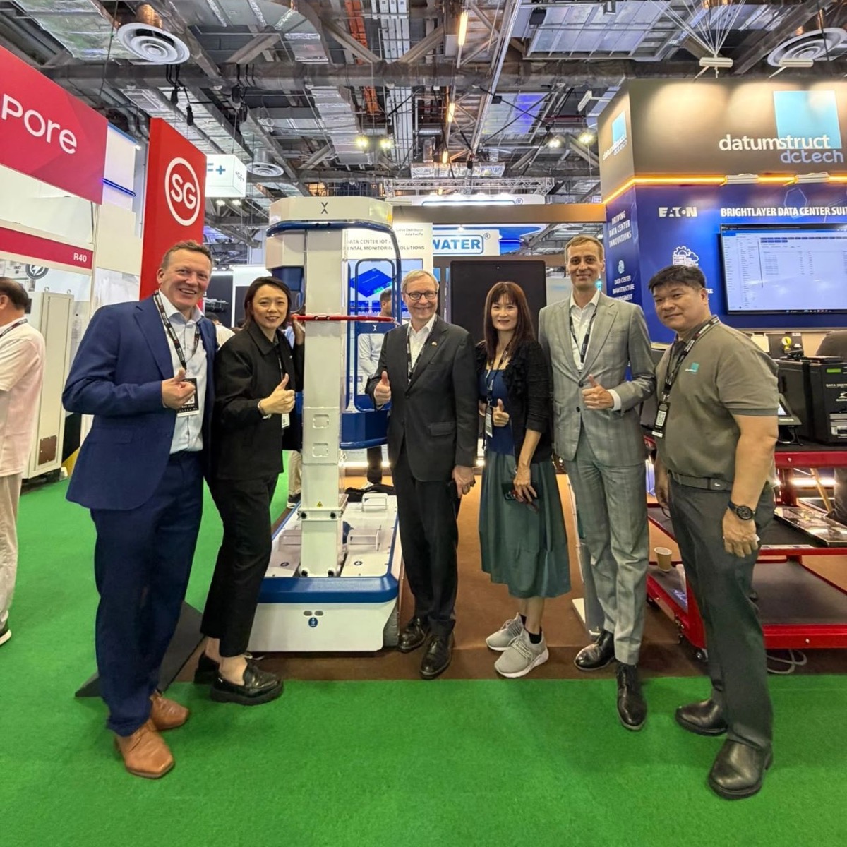 Six people on green carpet at the Datumstruct DC Tech booth at DCWA Singapore, most giving a thumbs up, around a white and blue IXOLIFT lifter